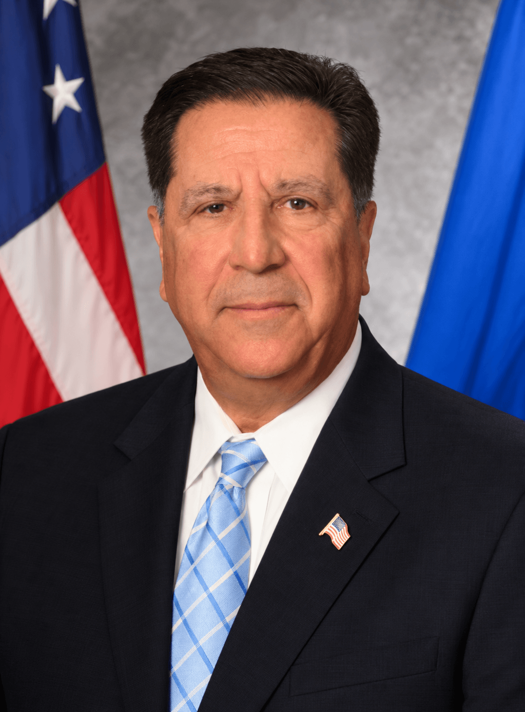 Man in black suit with light blue plaid tie standing before American and blue flags