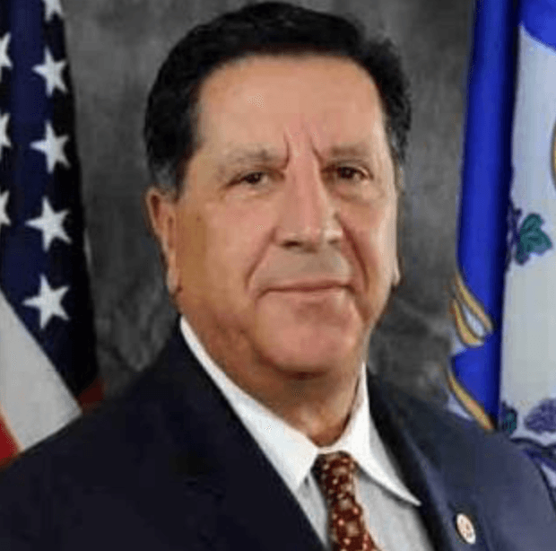 Professional headshot of man in dark suit and red patterned tie with American flag and state flag in background